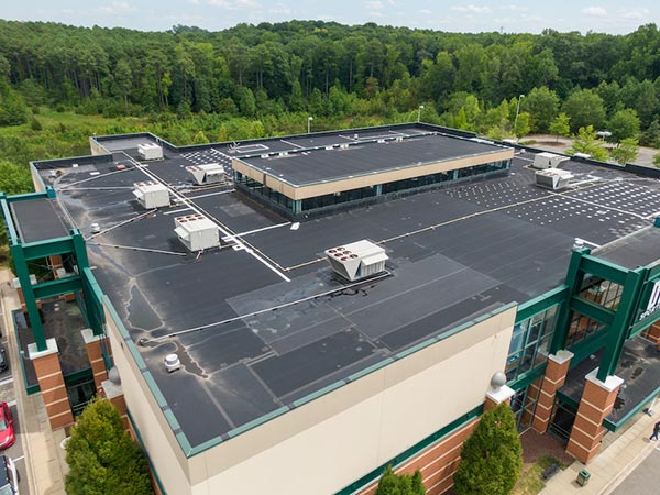 EPDM-Roofing-Onalaska-WI-Wisconsin-1 | Golden Eagle Roofing Aerial view of a large flat commercial roof with several white HVAC units and vents, bordered by a green-framed building and surrounded by trees.