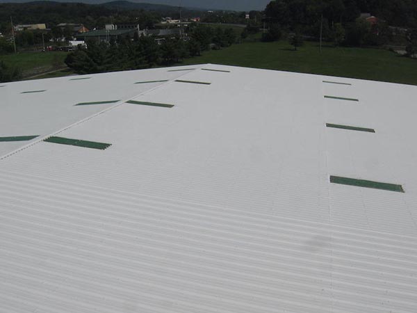 Aerial view of a large white corrugated metal roof with several rectangular green patches, with a grassy area and distant trees and buildings in the background.